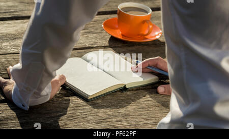 Rear view close-up of the hands of a man ready to write in front of an open notebook with blank pages next to a coffee cup. Stock Photo
