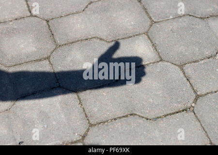 Close-up shot of shadow silhouette of human arm and hand with thumb-up success gesture on gray pavement lit by bright sun. Stock Photo