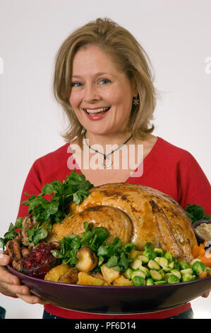 A roasted Christmas turkey with all the trimmings, Brussels sprouts, parsnips, bread sauce, cranberry sauce etc, being served by a blonde woman in red Stock Photo