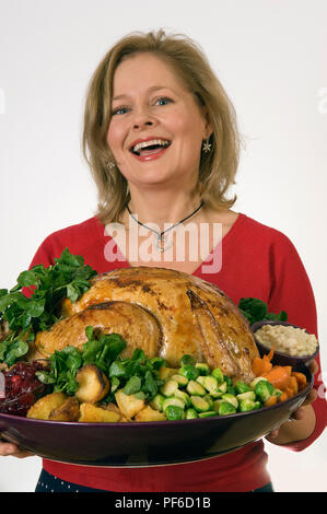 A roasted Christmas turkey with all the trimmings, Brussels sprouts, parsnips, bread sauce, cranberry sauce etc, being served by a blonde woman in red Stock Photo