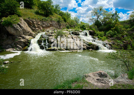 The waterfall on the river flows through and over the rocks covered with lichen and moss against a background of green vegetation and a blue sky. Stock Photo