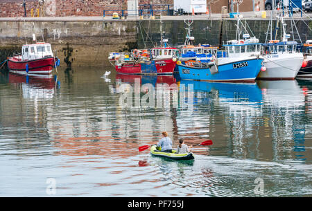 Dunbar, East Lothian, Scotland, United Kingdom, 20th August 2018. UK Sunshine in Dunbar harbour. Two women enjoy a paddle in an inflatable canoe among the colourful fishing boats moored in the harbour Stock Photo