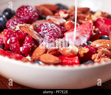 Homemade granola in a bowl, strawberries, almonds, blueberries, raspberry, honey ang pouring milk - ingredients for preparing natural dietary breakfas Stock Photo