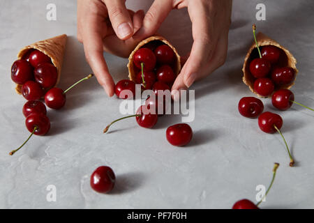 Three sweet waffle cones with ripe cherries holding girls hand on a gray background. Summer concept of homemade desserts. Stock Photo