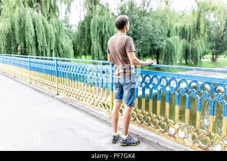 Young man leaning on bridge railing in green park in Myru or peace avenue in Naberezhna St, street, Rivne, western Ukraine looking at Ustya river in s Stock Photo