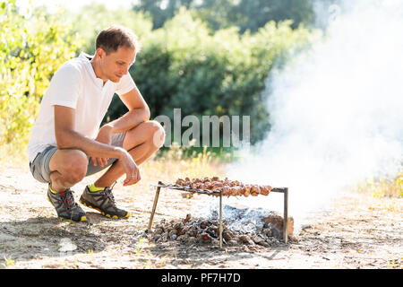 Young man grilling meat shashlik shish kebab skewers on barbeque bbq grill in outdoor, nature, summer park in Ukraine or Russia, sunny day, smoke Stock Photo