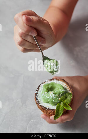 Woman's hands holding coconut with organic green ice cream on gray background. Stock Photo