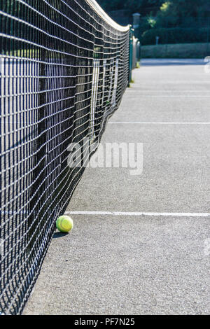 Low angle shot of of a yellow tennis ball on the ground next to the net of a hard surface tennis court Stock Photo