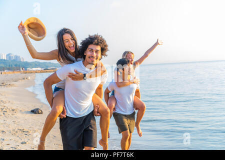 Group of friends walking along the beach, with men giving piggyback ride to girlfriends. Happy young friends enjoying a day at beach