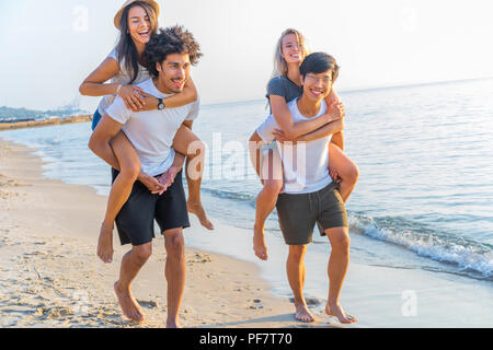 Group of friends walking along the beach, with men giving piggyback ride to girlfriends. Happy young friends enjoying a day at beach