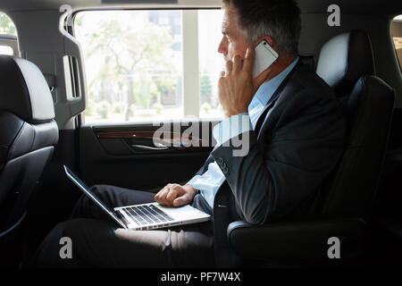 Handsome businessman talking with phone sitting with laptop on the backseat of the car. Stock Photo