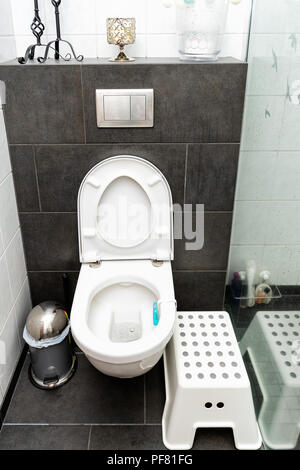 Clean modern toilet with open, opened cover, water, cleaner inside, trash rubbish can, assistance stool, chair, candles, vase and tiles, tiled floor i Stock Photo