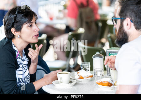 New Orleans, USA - April 23, 2018: Couple, man, woman sitting at table at famous, iconic Cafe Du Monde shop, drinking chicory coffee, eating beignet p