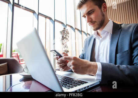 Businessman in black suit using mobile smart phone and working on laptop computer, browsing internet and writing on paper notebook in modern office. Man working on electronics devices with copy space Stock Photo