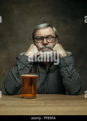 Sad bearded man drinking alcohol in pub. Enjoying my favorite beer. The front view of handsome smiling senior man with glass of beer sitting at the wooden table. Studio shot with caucasian model Stock Photo