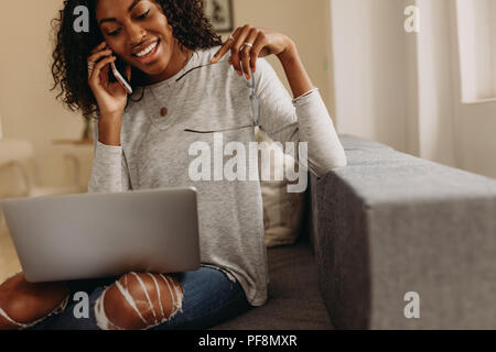 Woman entrepreneur in fashionable torn jeans speaking on mobile phone with laptop computer on her lap. Businesswoman sitting on sofa at home talking o Stock Photo