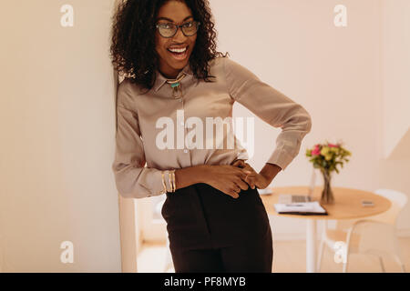 Portrait of a businesswoman in formal attire standing with hand on hip posing for photograph. Woman entrepreneur leaning on the wall sideways with lap Stock Photo