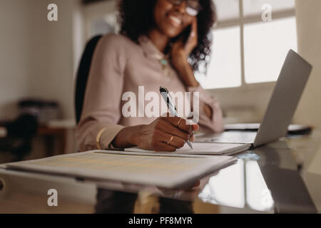 Businesswoman working on laptop computer and talking over mobile phone sitting at home. Close up of hand of a woman holding pen and writing on notepad Stock Photo