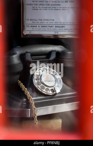 Close up of black vintage GPO bakelite telephone viewed through glass of disused red telephone box. Alpha numeric dial face, braided handset cord. Stock Photo