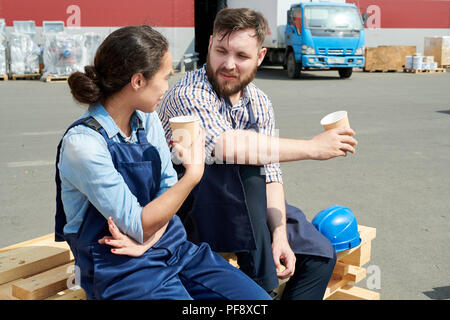 Portrait of two modern factory workers on break outdoors, drinking coffee and chatting sitting on stack of wood in sunlight Stock Photo