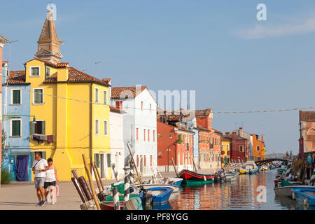Sunset in the colourful fishing village on Burano Island, Venice, Veneto, Italy. Canal scene with bright houses, reflections, boats and a couple walki Stock Photo