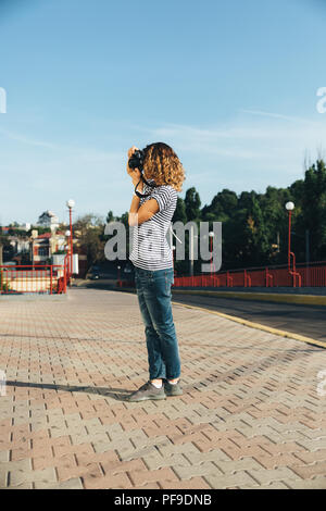 Vertical full body portrait of young woman in striped t-shirt and jeans taking photos. Side view female holding DSLR camera looking at viewfinder stan Stock Photo