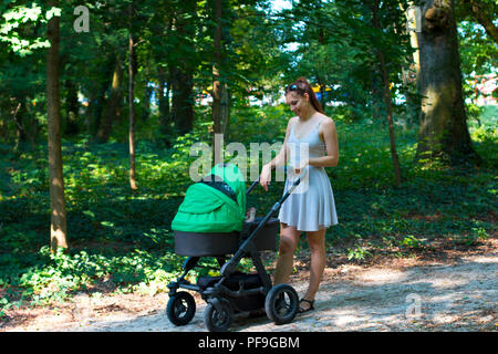 Nature walk with stroller, young mother in beautiful dress walking on the forest walkway with her baby in the pram, enjoying fresh air and smiling Stock Photo