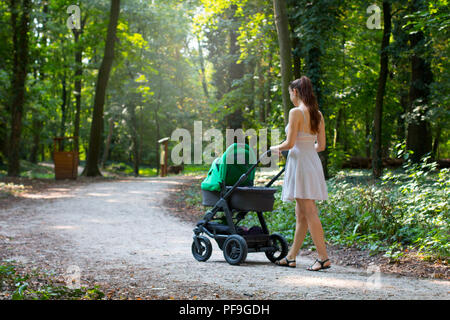 Woman in dress walking with the pram, surrounded by beautiful nature, pram walk on the walkway in the forrest park, parenting mother and baby outside Stock Photo