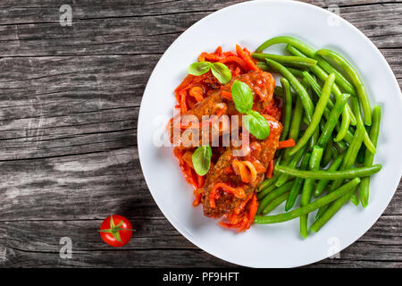 Delicious Meat Cutlets stewed with onion, carrot tomato sauce, decorated with basil leaves in a white dish with boiled green bean, top view Stock Photo