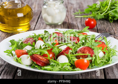 Salad with tomatoes, mozzarella, arugula, sausages on a white dish on a rustic table, balls of mozzarella cheese in a glass jar and bottle of olive oi Stock Photo