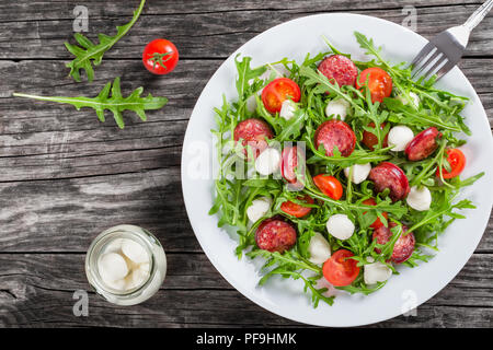 Salad with tomatoes, mozzarella, arugula, sausages on a white dish on a rustic table, balls of mozzarella cheese in a glass jar, blank space left, stu Stock Photo