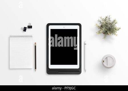 Tablet with blank screen for mockup on black case on white office desk. Notepad, pencil, clips, plant, plastic cup of coffee beside. Flat lay. Stock Photo