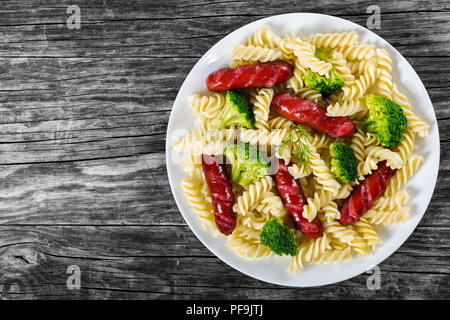 Delicious Spiral Pasta salad with  broccoli and grilled sausages decorated with dill on a white dish on an rustic table, studio lights, top view Stock Photo