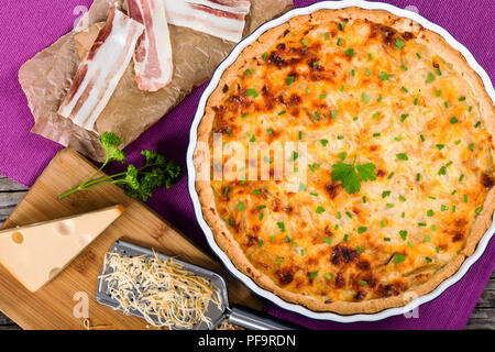 French onion quiche or pie in a gratin dish on table mat, with grater, piece of cheese on cutting board and slices of bacon on parchment paper,  view  Stock Photo