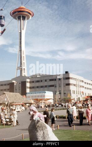 View of the attractions surrounding the Boulevards of World area of the Century 21 Exposition Seattle World's Fair, in Seattle, Washington, July, 1962. The Space Needle stands at left edge, behind the Food Circus building at center. Tucked into the Food Circus facade is the Behlen Building housing the Million Silver Dollar Display. Crowds of visitors walk among the fairground amusement rides and exhibits. At upper left edge is a Union 76 Sky Ride car traveling along its wires. At center foreground, a stuffed pink poodle souvenir toy is posed atop a large rock. () Stock Photo