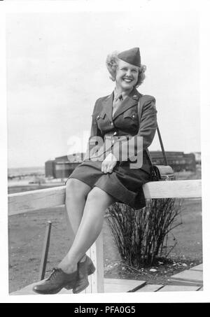 Black and white photograph, shot from a low angle, showing a smiling, blonde woman, in full length, wearing a military uniform and hat, with a handbag slung over one shoulder, sitting on a railing, outside on a sunny day, with a large building in the background, likely photographed in Ohio during World War II, 1945. () Stock Photo