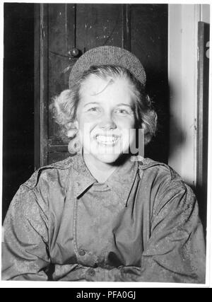 Black and white photograph showing a smiling young blonde woman, from the waist up, wearing a rain-soaked military jacket and a cap with the brim turned up, sitting in front of a cabinet, likely photographed during World War II, 1945. () Stock Photo