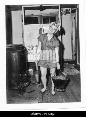 Black and white photograph, showing a blonde woman, in full-length view, facing the camera, wearing cut-off overalls, dirty sneakers, and a bandana tied around her hair, standing indoors next to a furnace, and holding a coal bucket and shovel, the open furnace appears to have a photograph shoved partially inside of it, likely photographed in Ohio during World War II, 1945. () Stock Photo