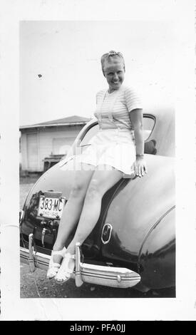 Black and white photograph, showing a smiling woman, in full length, perched on the back of a shiny, dark, vintage Chevrolet sedan car with an Ohio license plate dated 1945, wearing light colored shorts, a striped top, and a bandana wrapped around her hair, likely photographed in Ohio in the decade following World War II, 1950. () Stock Photo