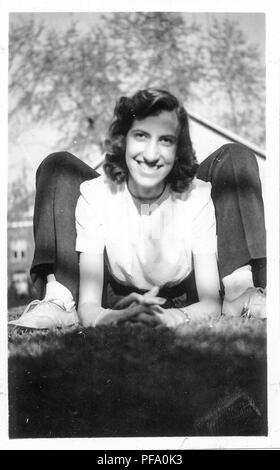 Black and white photograph, showing a smiling, dark-haired woman, outdoors, wearing a light colored top, seemingly contorted into a frog-like pose by lying on her stomach, and balancing on her elbows, while facing the camera, with a second person lying behind her on their back, with their legs visible immediately behind her shoulders, likely photographed in Ohio in the decade following World War II, 1950. () Stock Photo