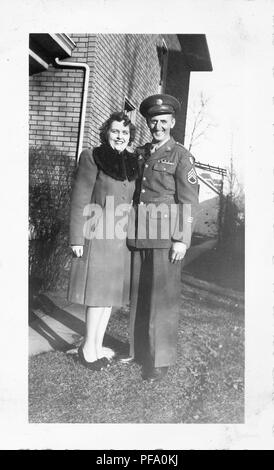 Black and white photograph, shot from a low angle, showing a smiling couple, both standing in full-length, and facing the camera, the man wearing a military uniform and cap, the dark-haired woman wearing bow-front, peep-toed heels and a knee-length winter coat with a dark fur collar, with a brick building visible in the background, likely photographed in Ohio during World War II, 1950. () Stock Photo