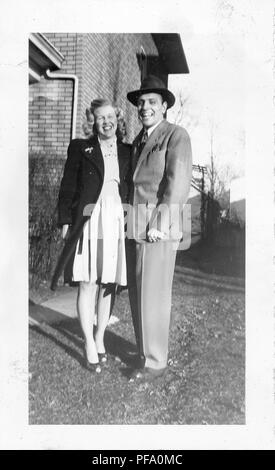 Black and white photograph, showing a full-length view of man standing outdoors, next to a blonde-haired woman, the woman wears a knee-length, light colored dress and a dark, knee-length coat with a brooch on the lapel, the man wears a suit, tie, and hat, both face the camera with broad smiles, likely photographed in Ohio in the decade following World War II, 1955. () Stock Photo