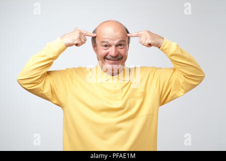portrait of a mature bald caucasian man pointing on temple with fingers ,an idea, dressed in a yellow pulover, isolated on a gray background. I want t Stock Photo