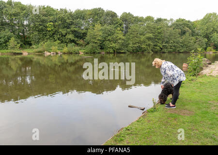 Mature blond lady stands at the side of the lake at Three Sisters Local Nature Reserve, Three Sisters Road, Ashton-In-Makerfield, Wigan with her black Stock Photo