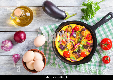 scrambled eggs, eggplant, onion and tomato in frying pan on kitchen towel,  garlic, aubergine,eggs,  olive oil,parsley and ripe tomatoes on wooden boa Stock Photo