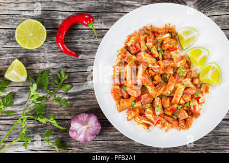 Braised Pig Ears or Oreja de Cerdo with spices, chili pepper, pieces of lime sprinkled with parsley on white platter on wooden old boards, top view Stock Photo
