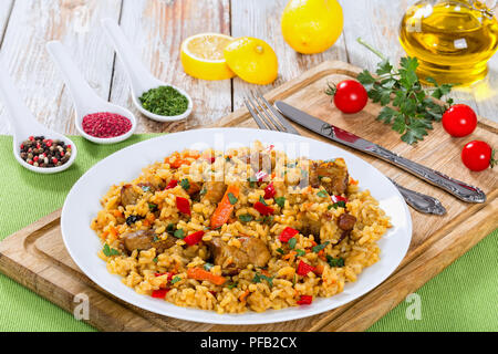 tasty paella with meat, pepper, vegetables and spices on platter on cutting board, lemon slice, spices and cherry tomatoes on background, view from ab Stock Photo