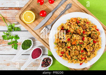 rice with meat, pepper, vegetables and spices on dish on cutting board, lemon slice, spices and cherry tomatoes on background, view from above Stock Photo