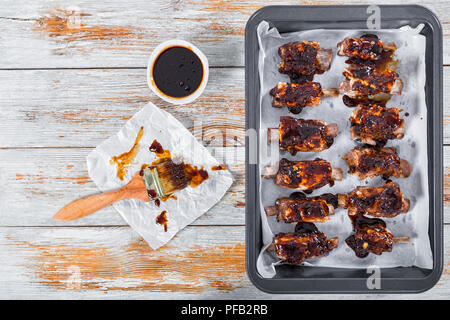 delicious sticky ribs seasoned with a spicy sauce prepared for grill in oven-tray on white peeling paint planks with basting brush, copy space left, v Stock Photo