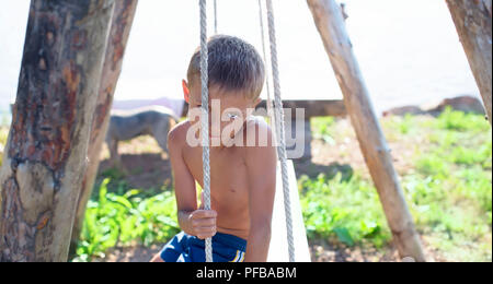 European boy sitting on a wooden old swing in a Park by the sea. Holidays in the village games in nature Stock Photo